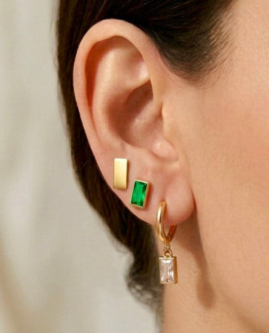 Close-up of a woman's ear wearing gold and emerald green earrings with a neutral background.